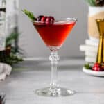 Side view of a Mistletoe Martini cocktail in a martini style glass on a gray countertop. The red colored cocktail is garnished with three cranberries on a gold cocktail pick and a sprig of fresh rosemary. To the right of the glass is a plate of cranberries and rosemary along with a gold jigger and a white and beige vase with evergreen sprigs. On the left are evergreen sprigs, a bottle of gin and a green bottle of vermouth, along with a beige and white linen.