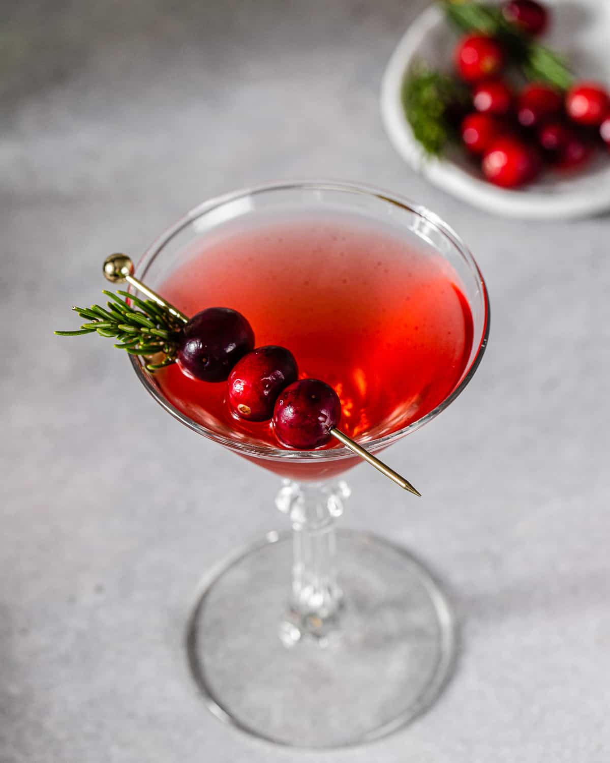 Overhead view of a Mistletoe Martini in a martini glass. There is a gold pick with three cranberries resting on top of the glass along with a sprig of rosemary. In the background on the top right is a plate of fresh cranberries and rosemary.