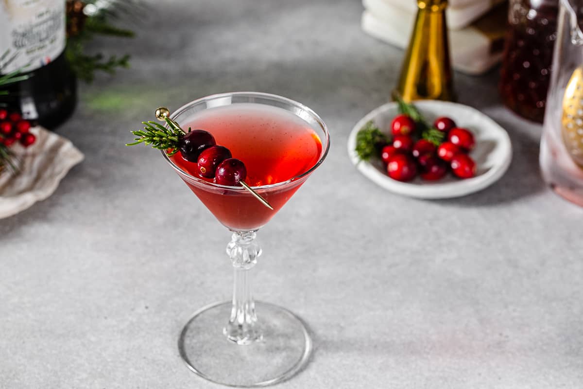 Slight overhead view of a Mistletoe Martini in a martini glass. There is a gold pick with three cranberries resting on top of the glass along with a sprig of rosemary. In the background on the top right is a plate of fresh cranberries and rosemary along with a gold jigger and some greenery. To the left is a beige and white striped linen and a bottle of dry vermouth.