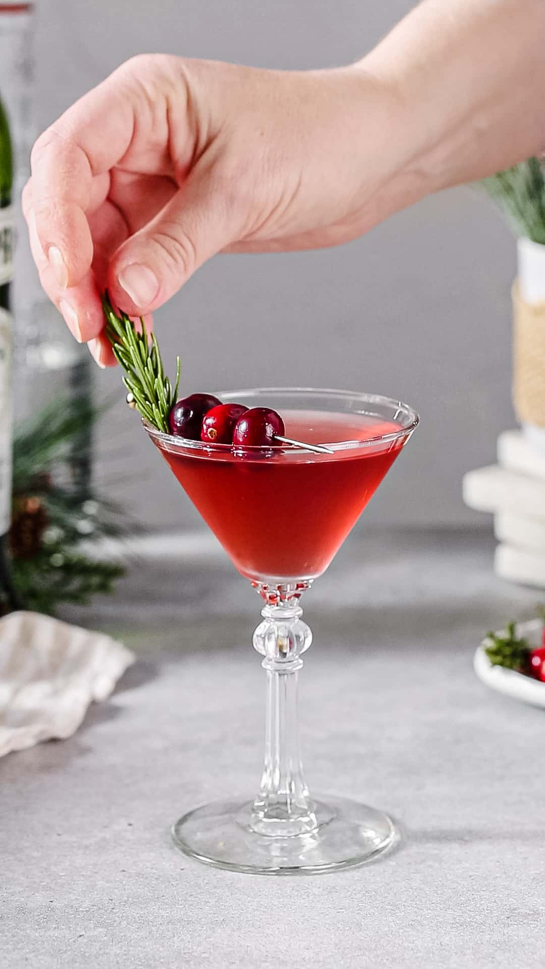 Hand adding a sprig of fresh rosemary to the top of a martini glass filled with red liquid and garnished with cranberries.