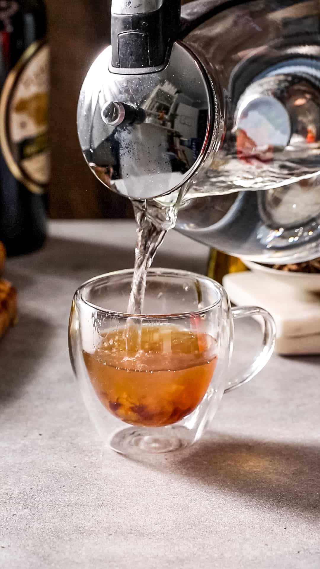 Hot water pouring from a kettle into a glass mug filled with an amber colored liquid.