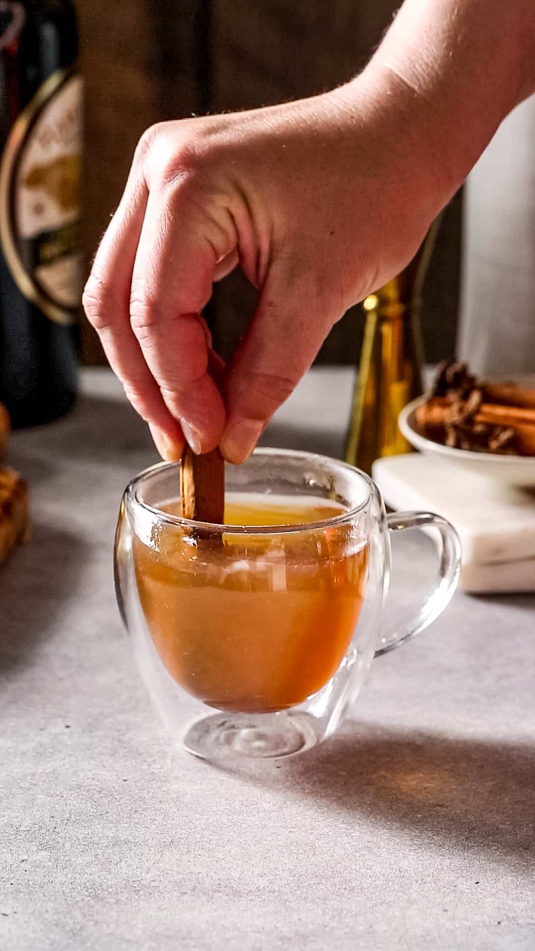 Hand using a cinnamon stick to stir amber liquid in a glass mug.