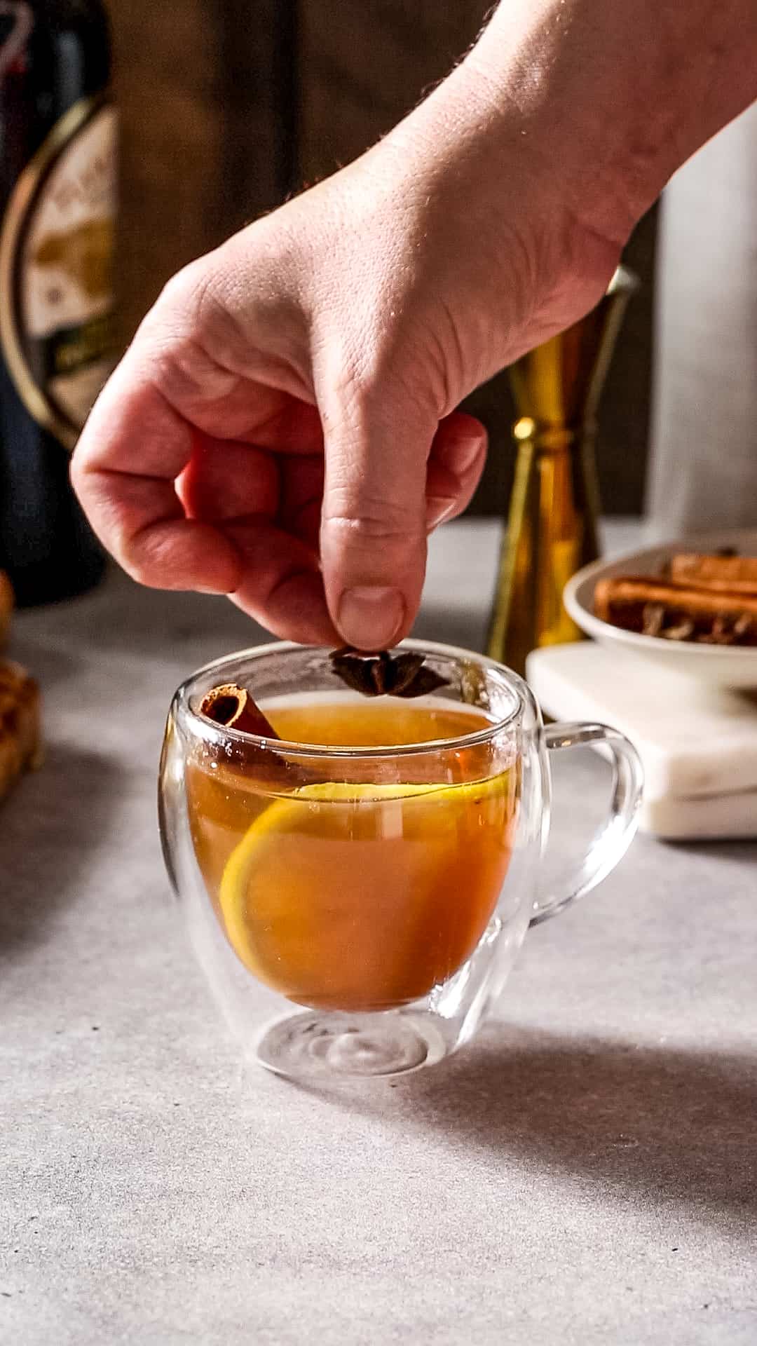 Hand adding a star anise to a glass mug filled with amber liquid, a cinnamon stick and a lemon slice.