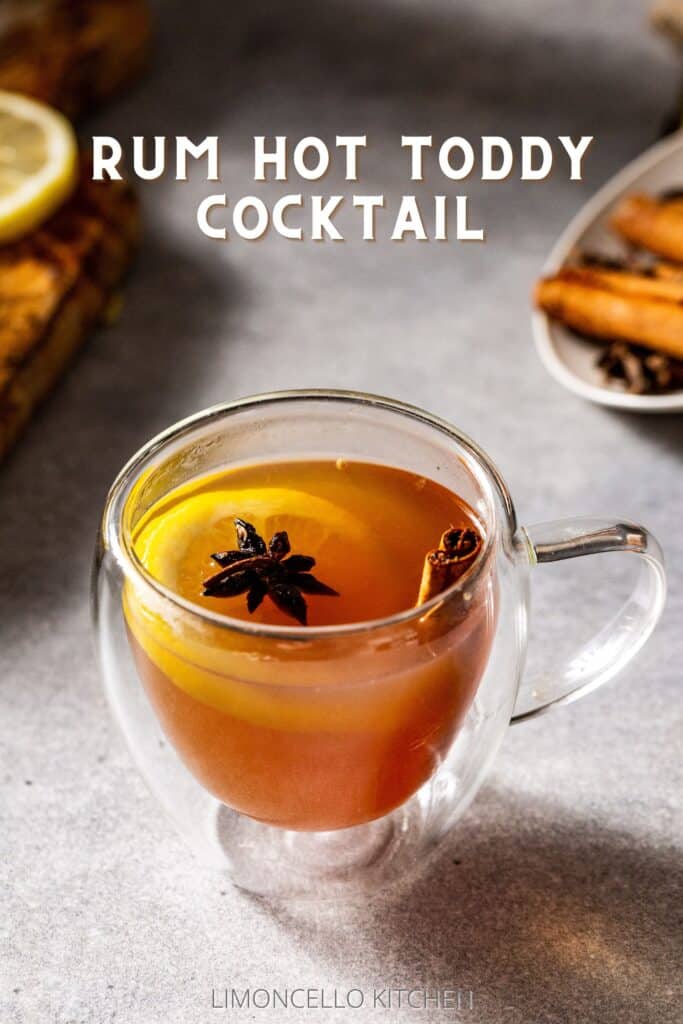 Overhead view of a Rum Hot Toddy cocktail in a glass mug on a gray countertop with a cinnamon stick, star anise and lemon slice in the glass as a garnish. Lemons are on a wooden cutting board on the left, and on the right is a dish of cinnamon and star anise. Text saying "Rum Hot Toddy Cocktail" is overlaid above the drink.