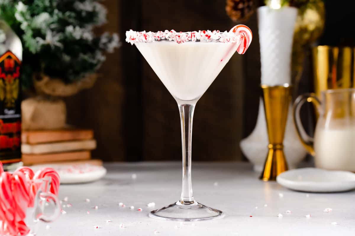 Side view of a Candy Cane Martini in a martini style glass. The drink is white and the rim of the glass is coated in crushed candy cane pieces. A mini candy cane is on the rim of the glass. To the left of the drink, mini candy canes are in a jar, and in the background there is a bottle of peppermint schnapps and some evergreen decor. To the right of the drink are a jigger, small pitcher of milk, a white plate and vase, and a gold cocktail shaker.