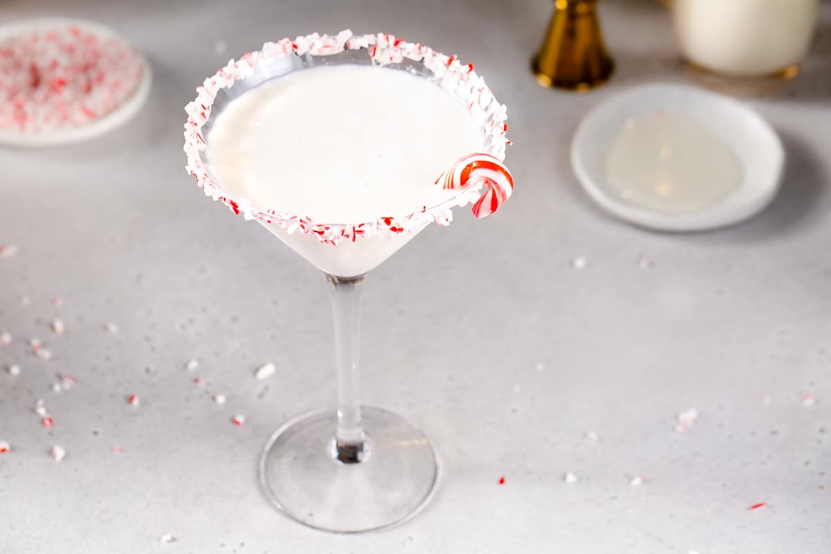 Overhead view of a Candy Cane Martini in a stemmed martini style glass. The drink is white and the rim of the glass is coated in crushed candy cane pieces. A mini candy cane is on the rim of the glass. It is sitting on a gray countertop along with a dish of crushed candy canes, a gold jigger and a dish of corn syrup.