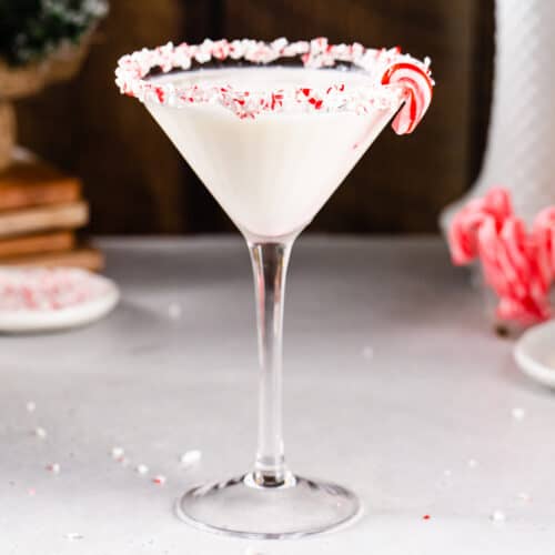 Side view of a Candy Cane Martini in a martini style glass. The drink is white and the rim of the glass is coated in crushed candy cane pieces. A mini candy cane is on the rim of the glass. To the left, a dish of crushed candy canes and some wooden coasters are visible. To the right, a white plate, a small glass filled with mini candy canes, and a white vase are visible.
