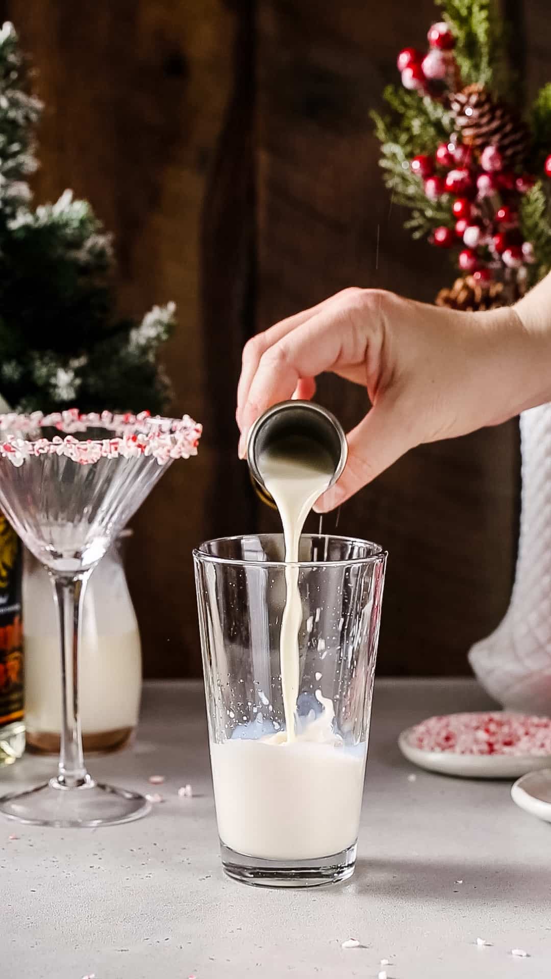 Hand pouring milk from a jigger into a glass cocktail shaker.