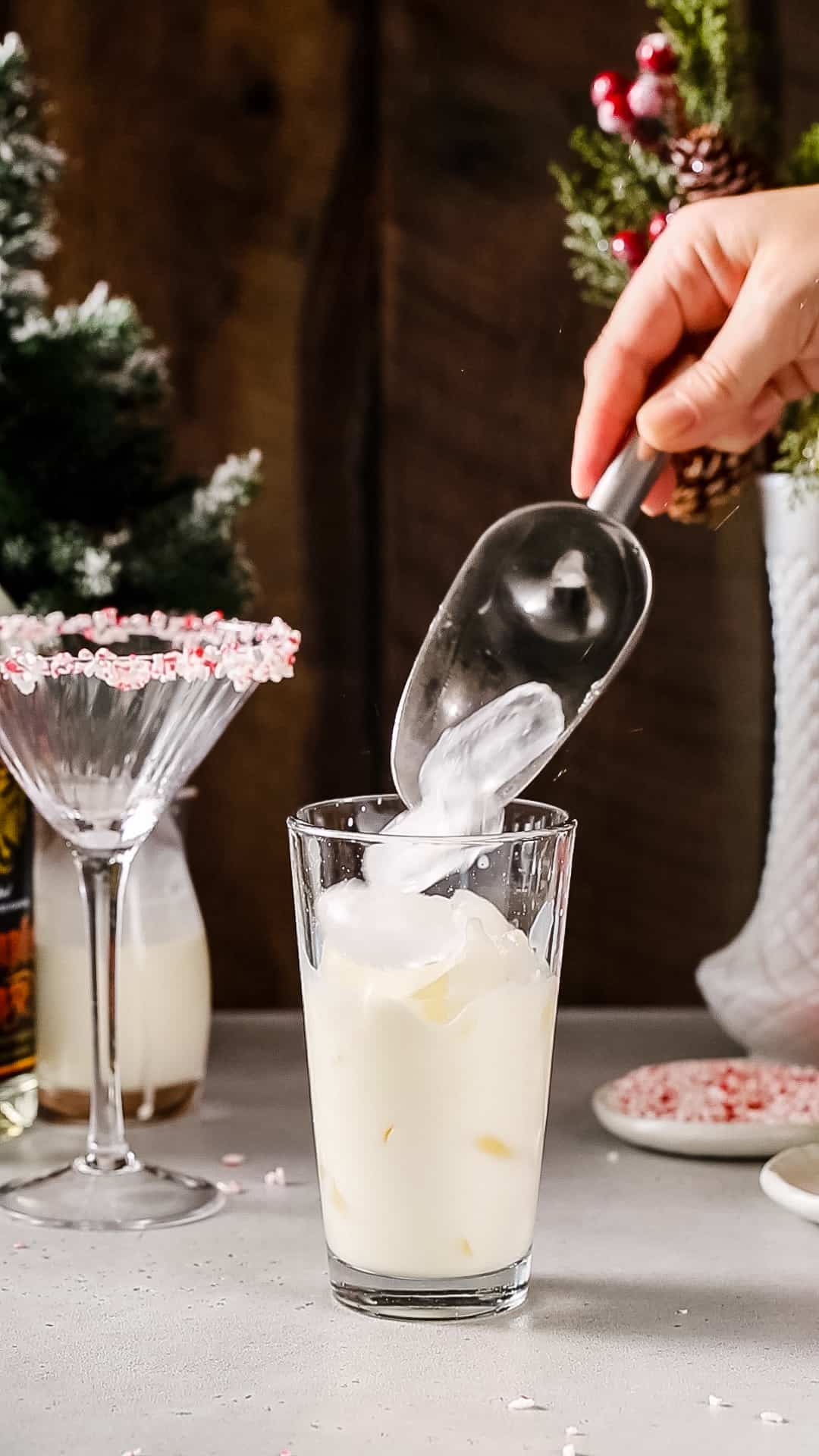 Hand using an ice scoop to add ice to a glass cocktail shaker filled with white creamy liquid.