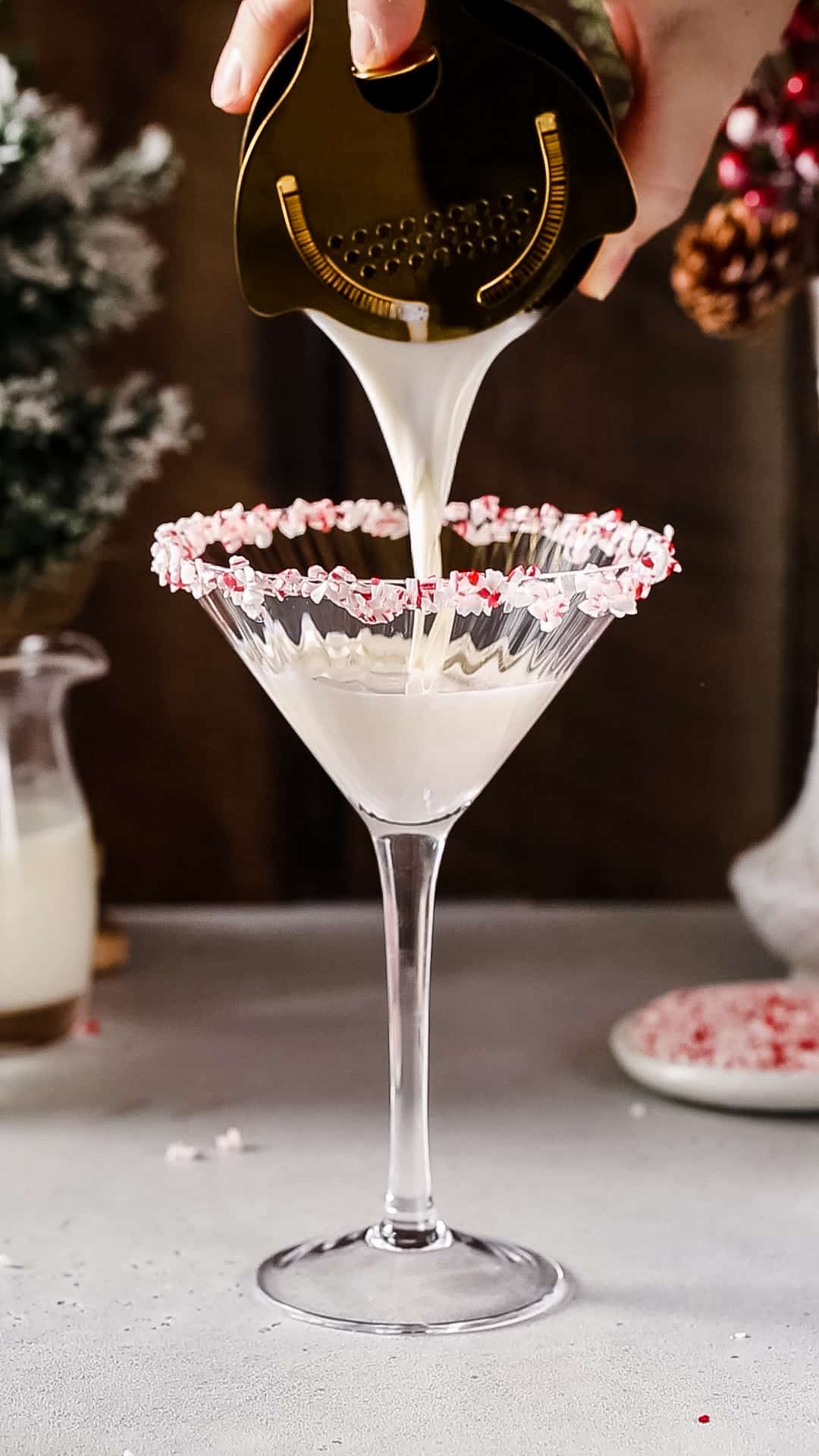 Hand pouring a white cocktail into a martini glass with a crushed candy cane rim.