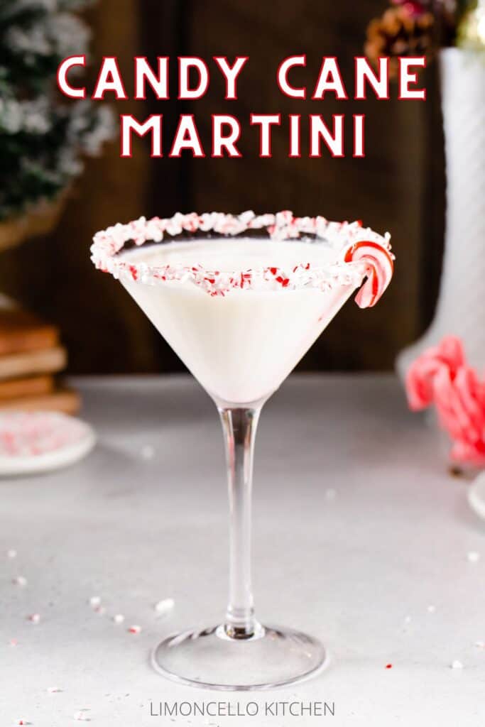Side view of a Candy Cane Martini in a martini style glass. The drink is white and the rim of the glass is coated in crushed candy cane pieces. A mini candy cane is on the rim of the glass. To the left, a dish of crushed candy canes and some evergreen decor are visible. To the right, a white plate, a small glass filled with mini candy canes, and a white vase are visible. Text above the drink reads “Candy Cane Martini” in all capital letters, in white color with a red outline. Smaller text below the drink reads “Limoncello Kitchen”.