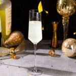 Side view of an Italian 75 cocktail in a Champagne flute. A bottle of Limoncello is visible in the background along with a gold jigger and gold decor. The drink is garnished with a fancy lemon peel on the rim.