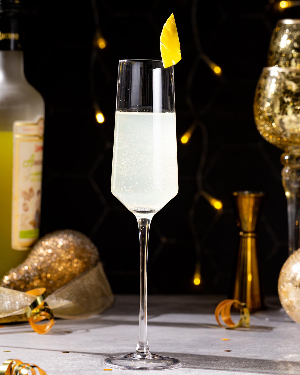 Side view of an Italian 75 cocktail in a Champagne flute. A bottle of Limoncello is visible in the background along with a gold jigger and gold decor. The drink is garnished with a fancy lemon peel on the rim. Gold ribbon curls and gold confetti are on the countertop.