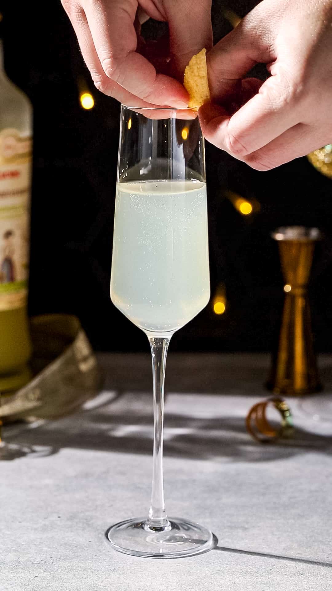 Hands adding a lemon peel twist garnish to the rim of a Champagne flute filled with sparkling yellow liquid.
