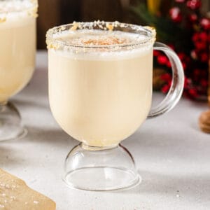 Close up of a glass mug filled with a Sugar Cookie Eggnog cocktail. The rim is coated in sugar cookie crumbs and there is a sprinkling of nutmeg on top. Another mug sits slightly behind it and some sugar cookies are visible to the bottom left. Red and green decor is in the background.