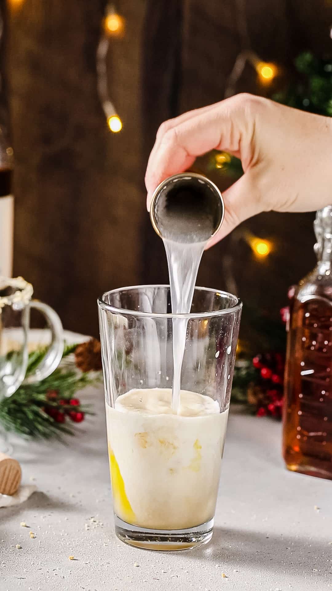 Hand pouring vanilla vodka from a jigger into a cocktail shaker filled with creamy liquid.