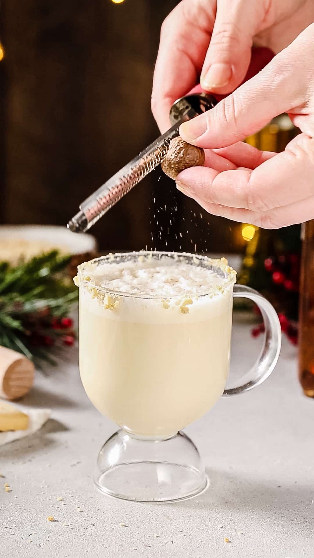 Hands using a spice grater to grate fresh nutmeg over top of a mug of eggnog.