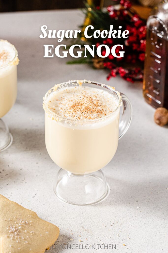 Slightly overhead shot of two mugs of Sugar Cookie Eggnog on a gray countertop. A few Christmas tree-shaped sugar cookies are on the left. On the right is a bottle of sugar cookie syrup and some evergreen and berry decor, and a gold cocktail jigger. Overlay text at the top reads “Sugar Cookie Eggnog”.