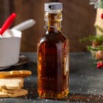 A bottle of Sugar Cookie Syrup on a countertop with sugar cookies to the left of the bottle. The bottle is capped with a grey colored cap. In the background are a white pot with a red spatula in it and some evergreen and berry holiday decorations.