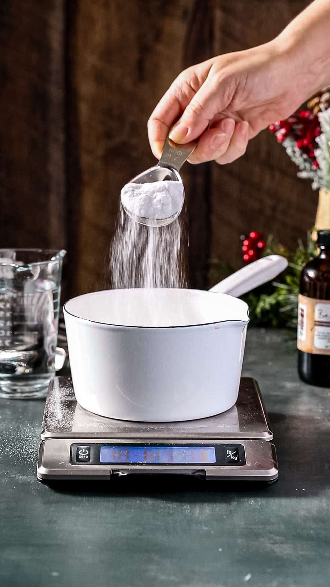 A hand adding sugar to a white pot on a baking scale.