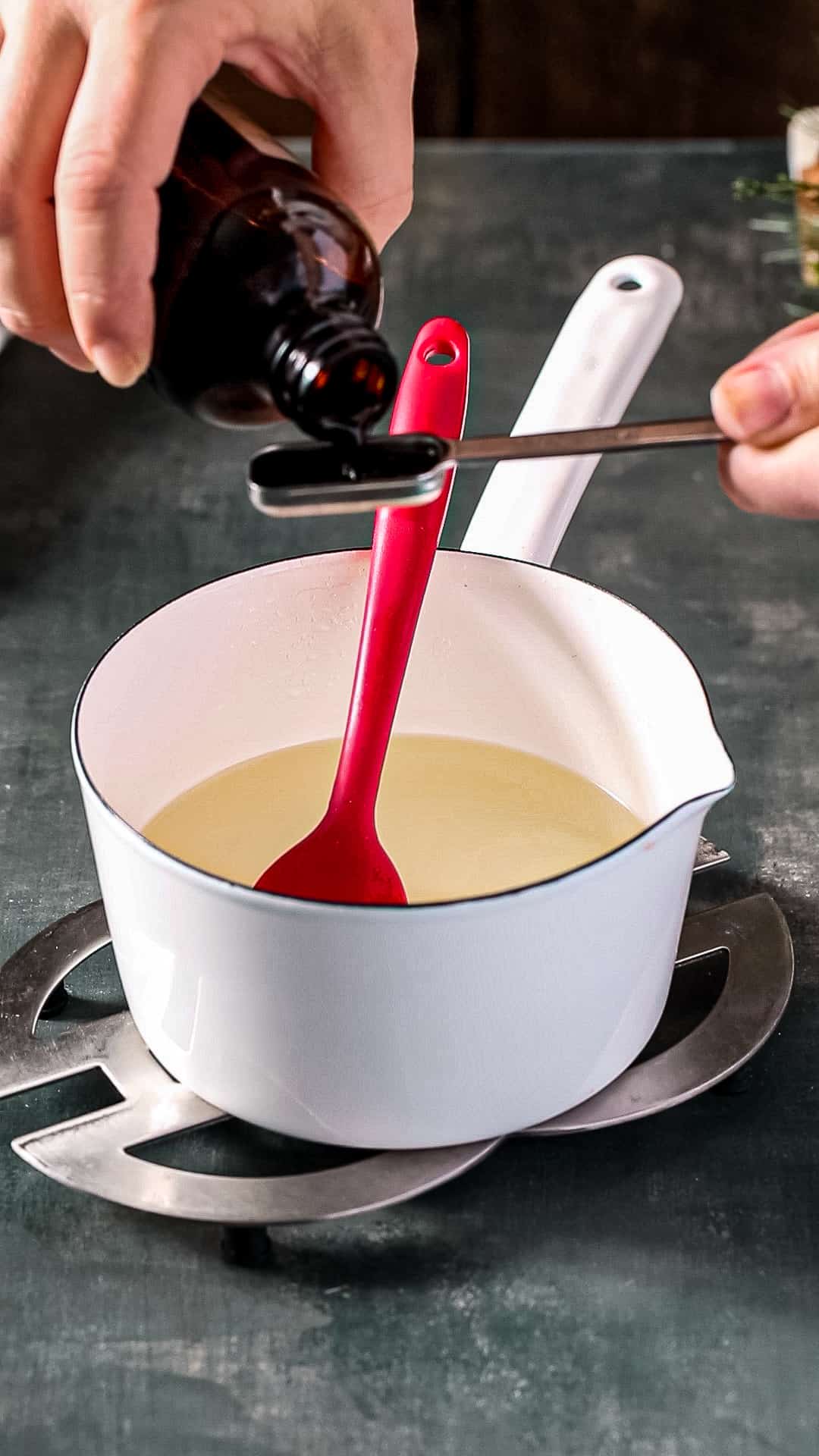 Hand measuring out vanilla extract over top of a white pot with liquid inside.