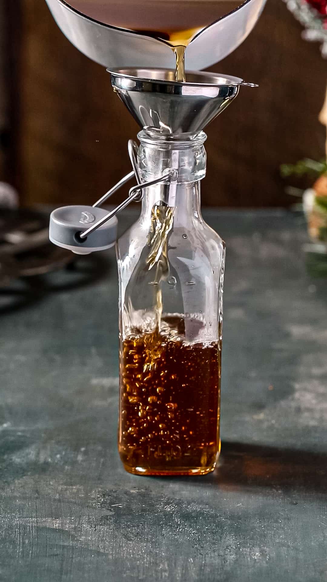 Brown colored syrup being poured through a funnel sitting on top of a swing top bottle. The bottle is halfway filled in this action shot.
