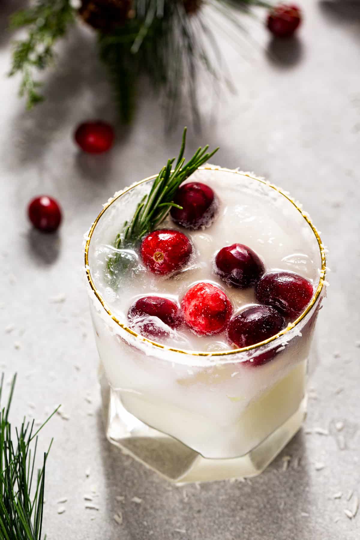 Overhead shot of a White Christmas Margarita, a white colored drink with cranberries and rosemary garnish with a dried coconut rim. The drink is surrounded by fresh cranberries and evergreen sprigs.
