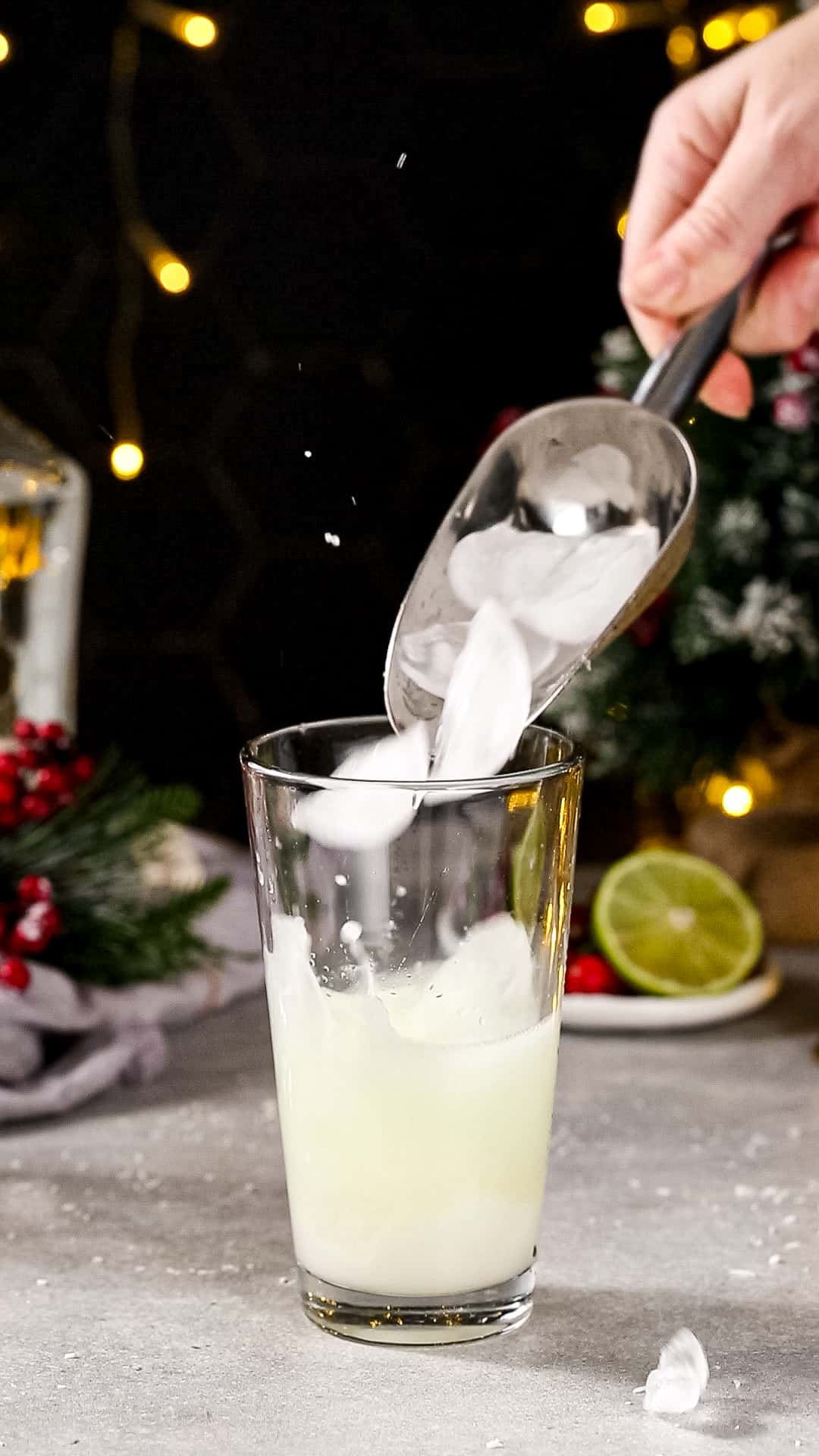 Hand using a silver ice scoop to add ice to a glass cocktail shaker filled with white liquid.