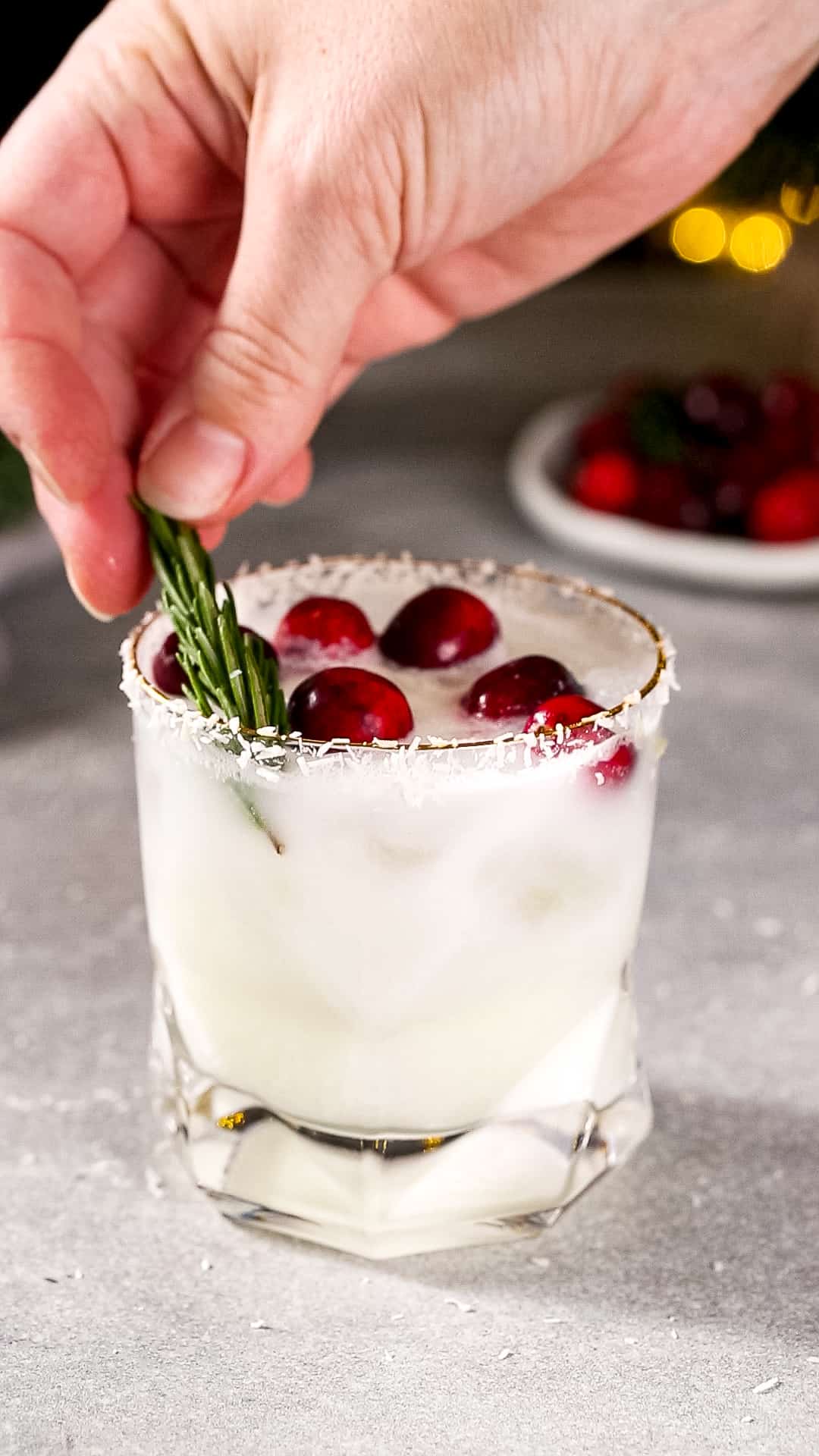 Hand adding a sprig of rosemary to a white drink in an old fashioned glass.