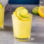 Close up of a Citrus Slush mocktail on a gray countertop with a lemon and lime slice twisted on top as a garnish. Another drink is off to the left, and some cut up citrus fruit is in the background on the right.