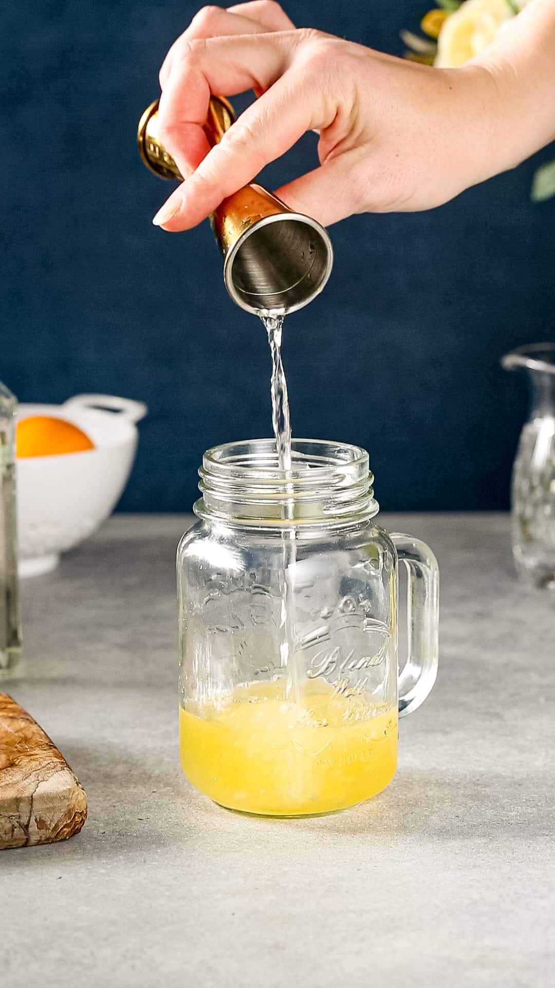 Hand using a gold jigger to pour water into a mason jar that has orange colored liquid in it.