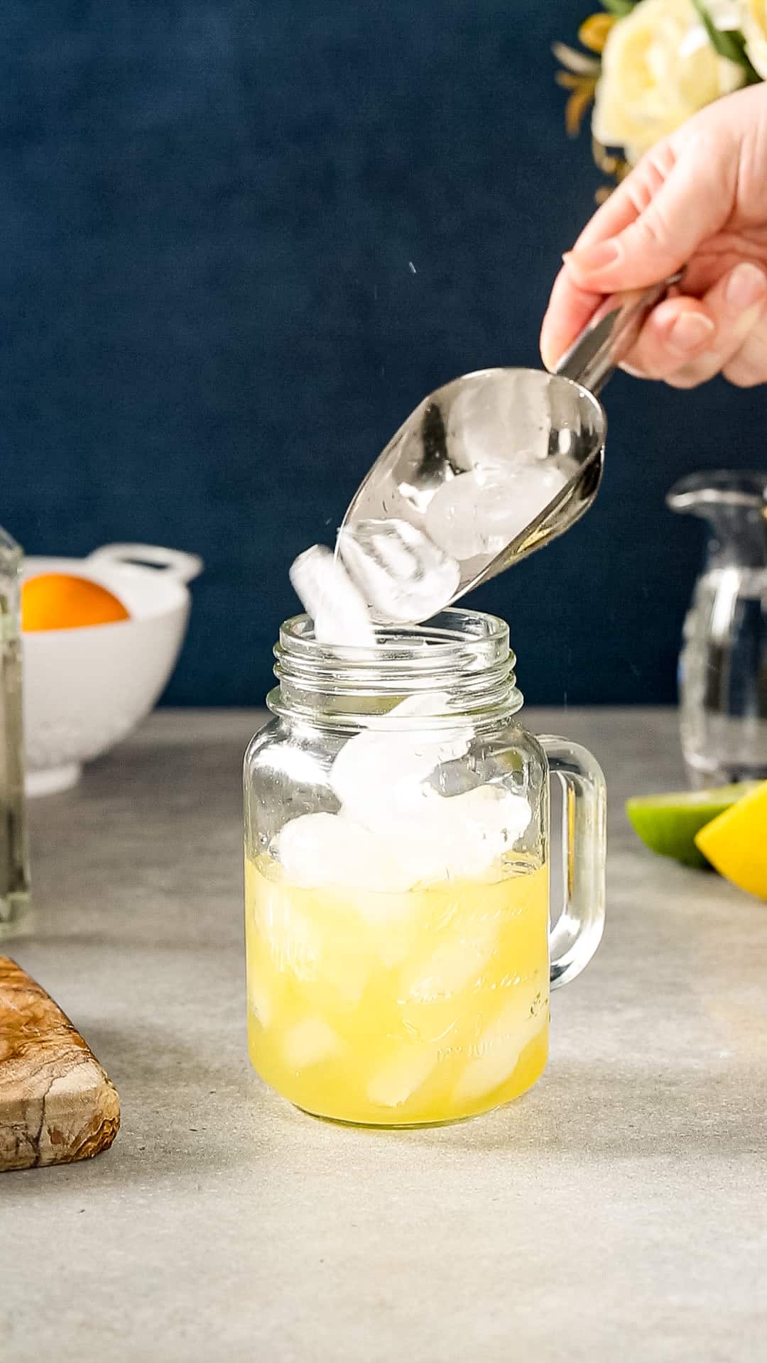 Hand using a silver ice scoop to fill a mason jar with ice. The mason jar also has yellowish orange liquid in it.