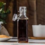 Side view of an open swing-top glass bottle filled with dark brown Demerara syrup on a gray countertop. A small dish of demerara sugar is to the left of the bottle. In the background, a white saucepan is on the right on top of a cork trivet, and a tan colored linen is on the left along with some greenery. There is a dark brown wood wall in the back.