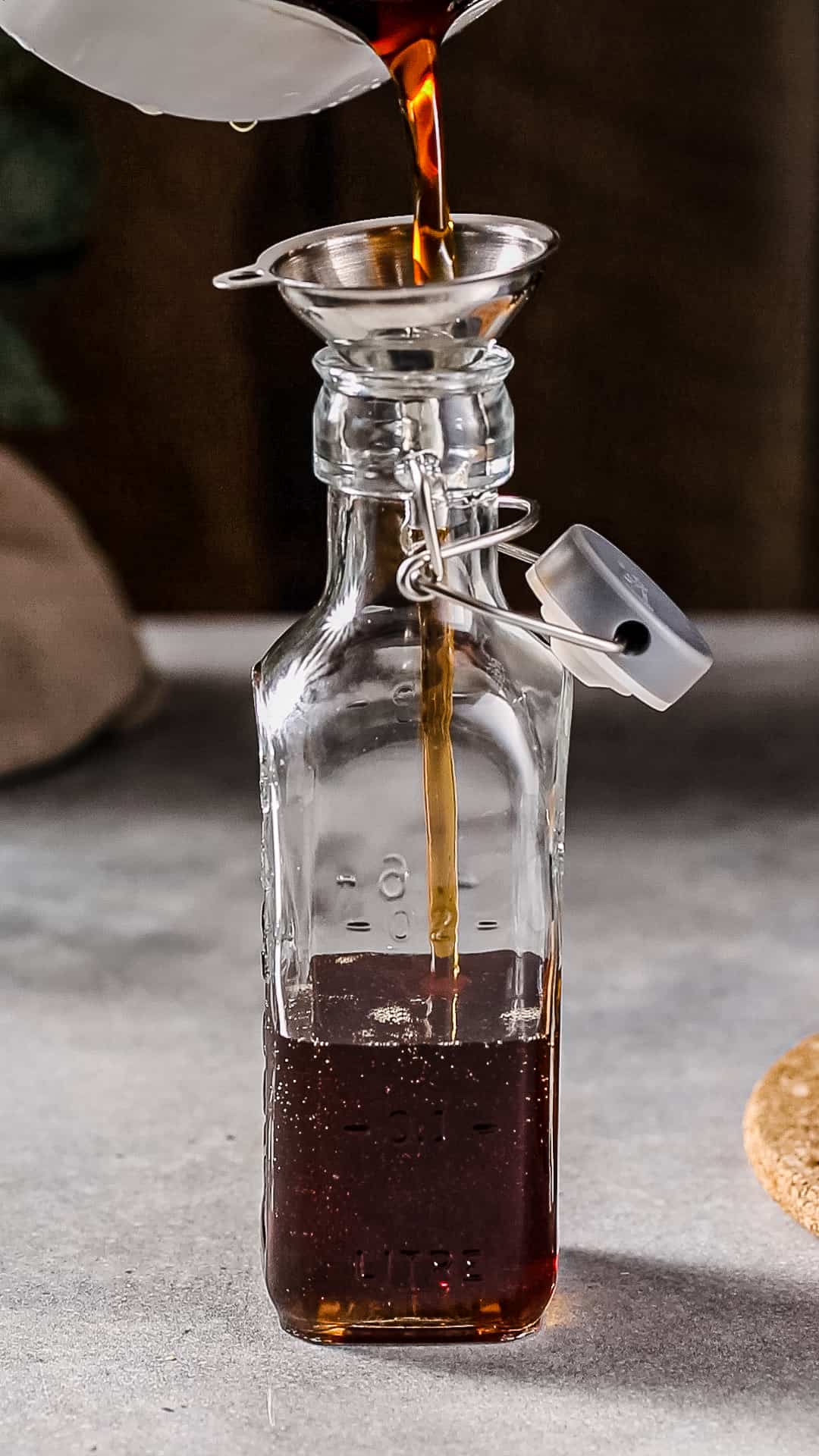 Brown liquid being poured from a white saucepan through a funnel into a glass swing-top bottle.