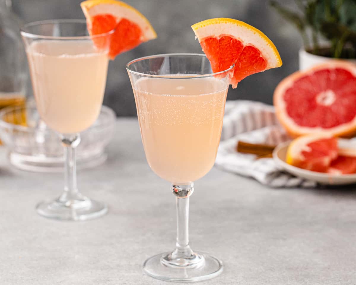 Grapefruit Mocktail in a goblet style wine glass on a gray countertop, with a quarter slice of grapefruit as a garnish on the rim of the glass. Cut fresh pink grapefruit is in the background on the right along with a gray striped linen. On the left is another mocktail.