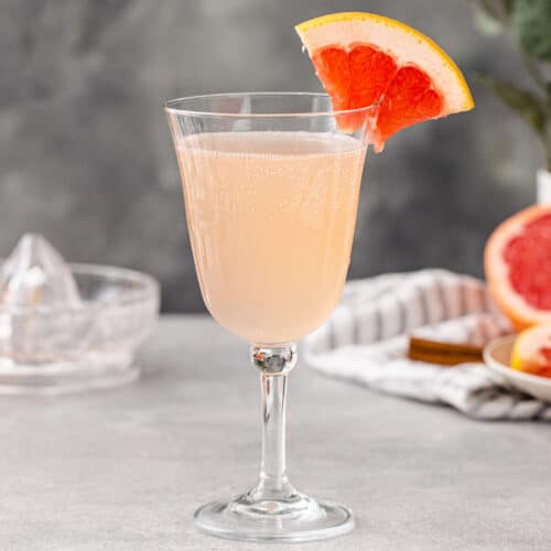 Side view of a Grapefruit Mocktail in a stemmed wine glass with a flared rim. There is a quarter slice of a pink grapefruit as a garnish on the rim of the glass. Cut fresh pink grapefruit is in the background on the right along with a gray striped linen and some cinnamon sticks.