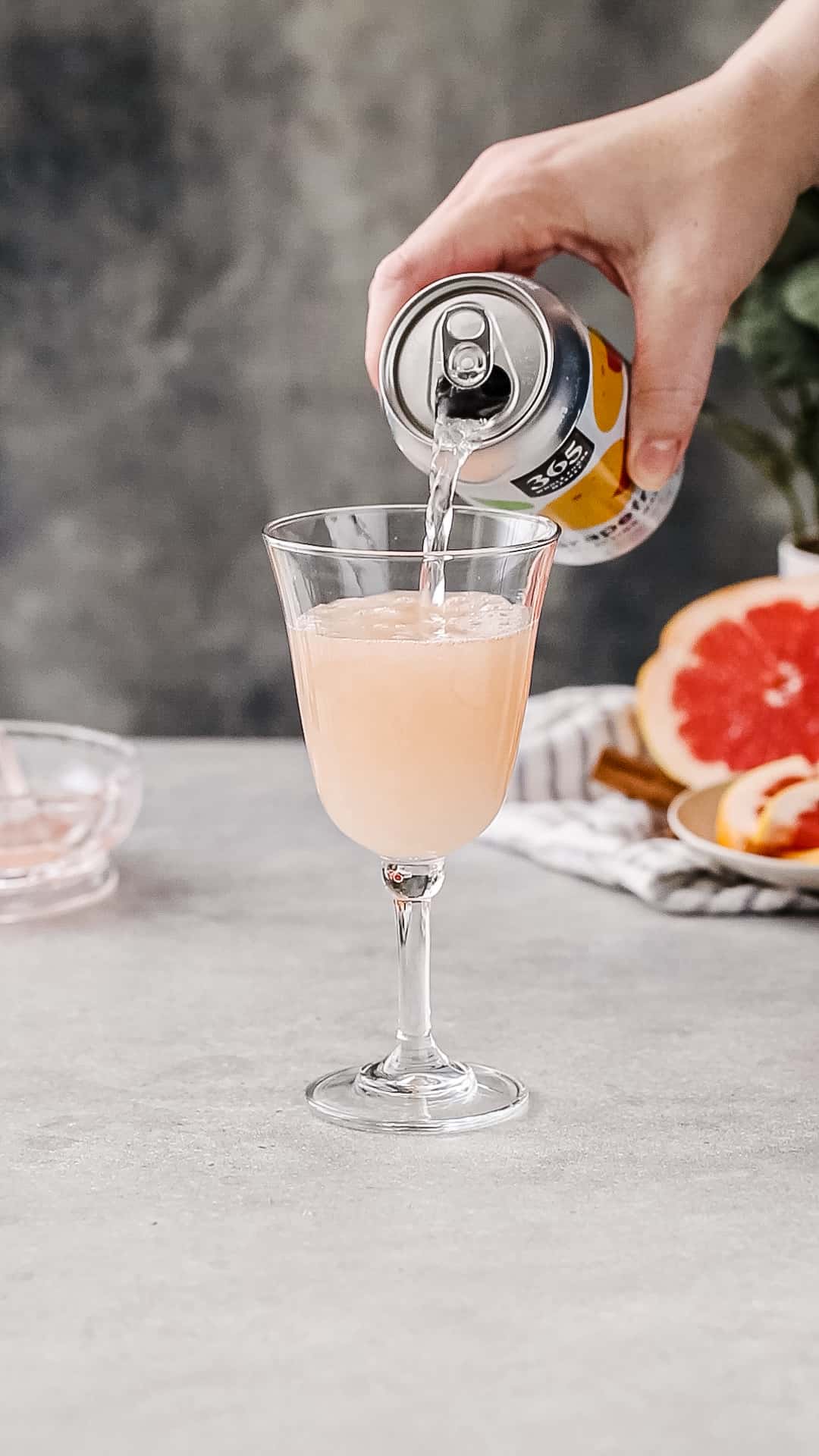 Hand pouring grapefruit seltzer from a can into a cocktail serving glass filled with pink liquid.