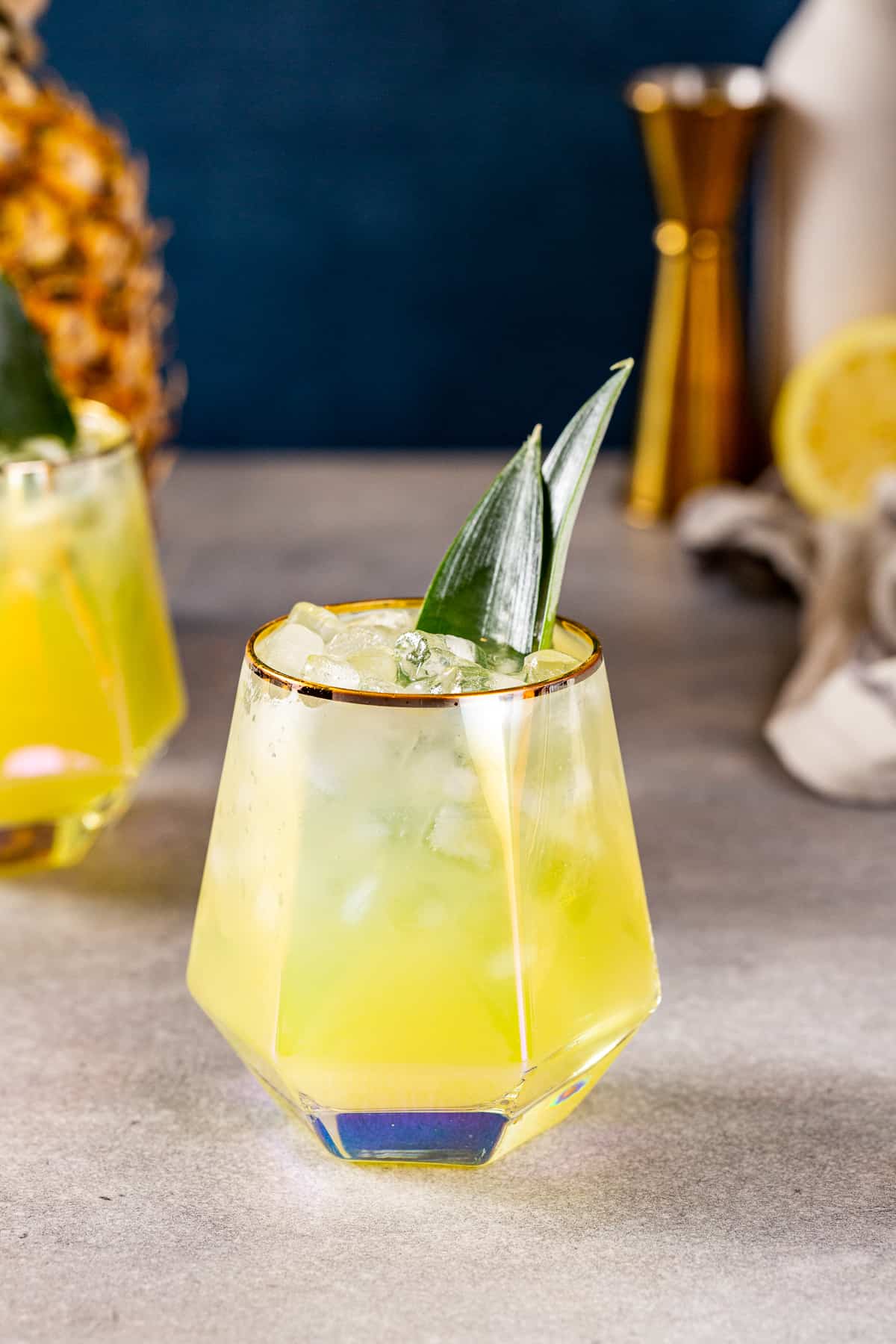 Pineapple mocktail on a countertop with another drink in the background. Also visible in the background is a pineapple on the left and a cut lemon and a gold jigger on the right.