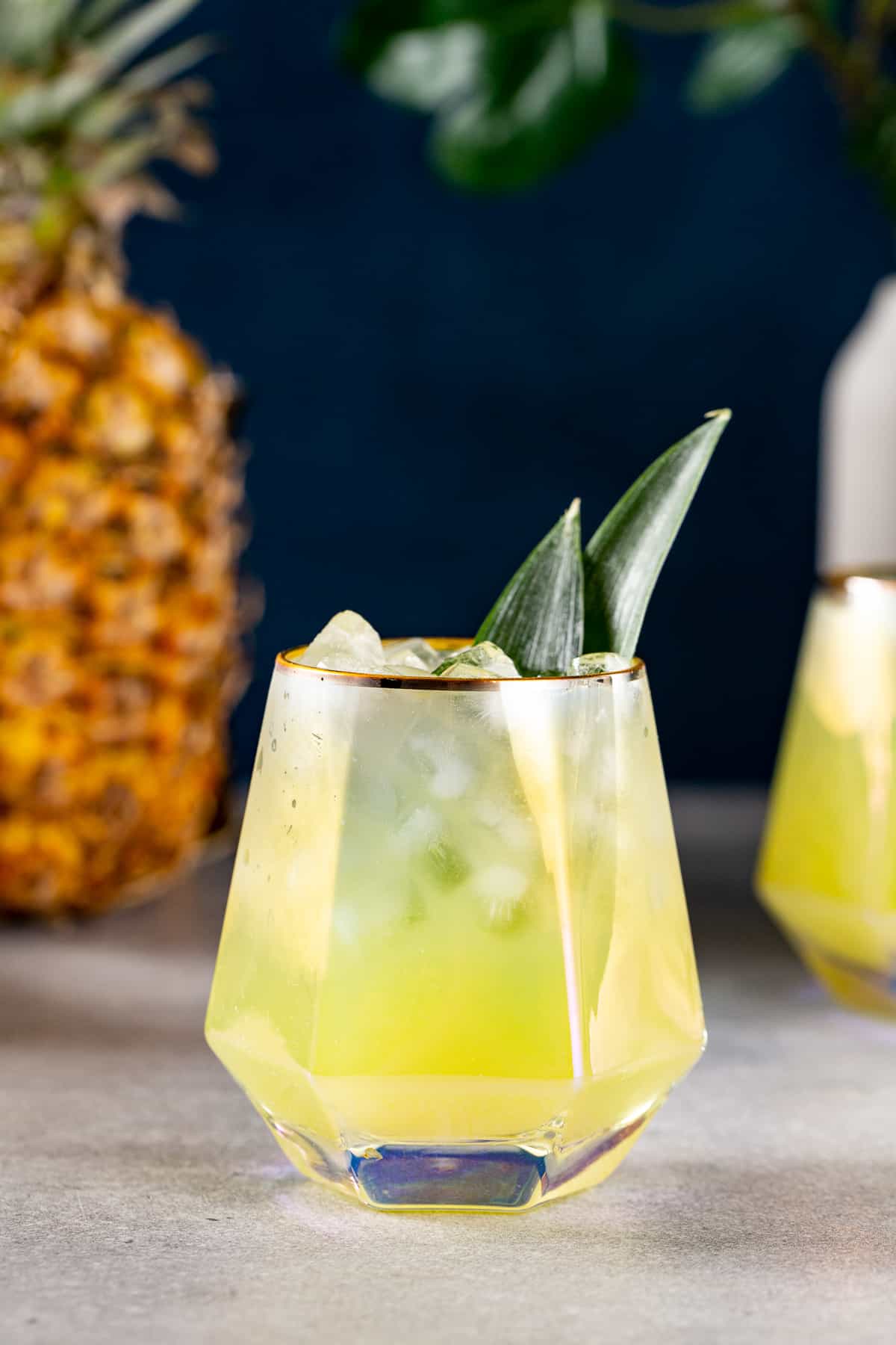 Side view of a pineapple mocktail in a geometric cocktail glass with pineapple fronds as a garnish. A whole pineapple is visible on the left.