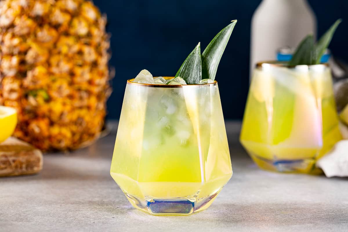 Side view of a pineapple mocktail in a geometric cocktail glass with pineapple fronds as a garnish. A whole pineapple is visible on the left and another drink is off to the right.