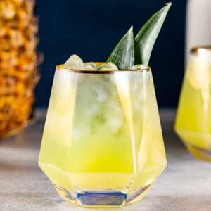 Close up of a pineapple mocktail in a geometric glass with pineapple frond garnish. A whole pineapple is off to the left.
