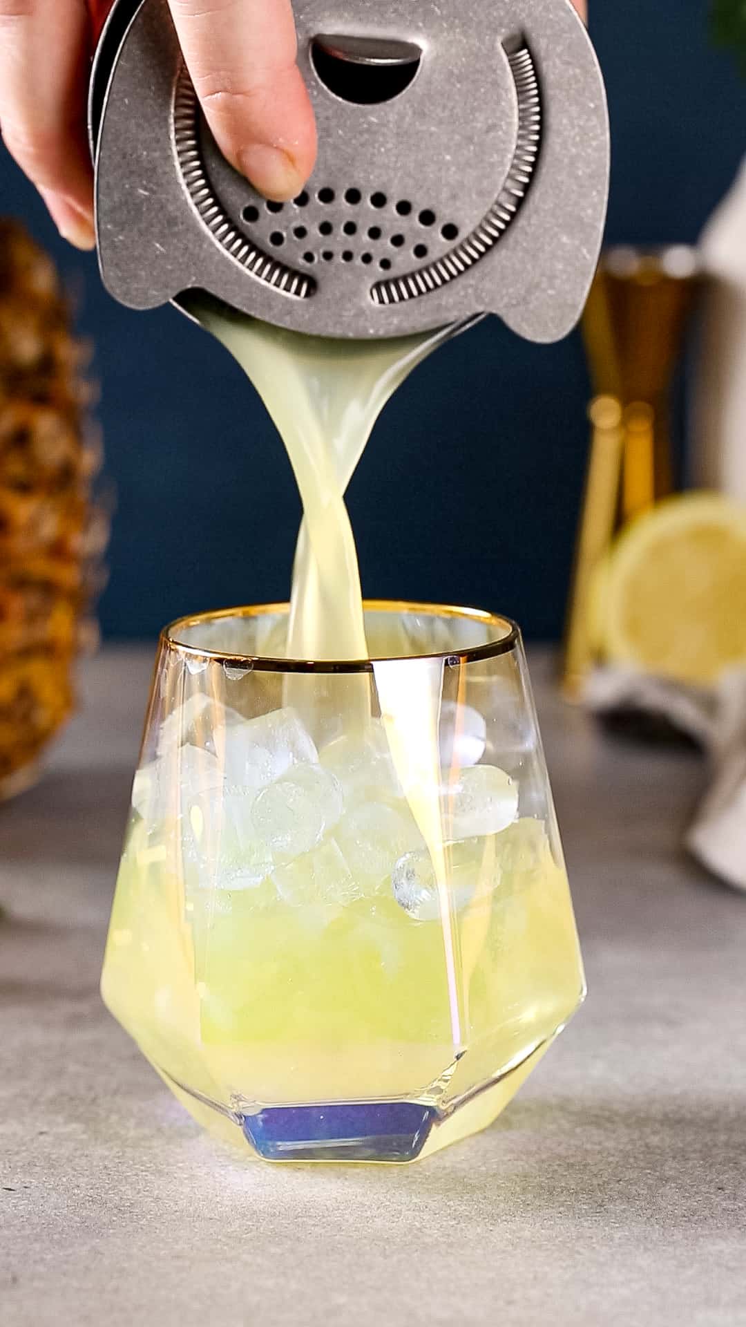Hand using a cocktail shaker to strain a yellow drink into a serving glass.