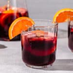 Side view of non-alcoholic sangria mocktail in a lowball glass. The drink is deep red with fruit slices floating in it and an orange half moon garnish on the rim of the glass. In the background is a second mocktail on the right and a half full glass pitcher on the left.