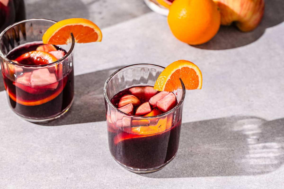 Overhead view of non-alcoholic sangria in low ball glasses on a gray countertop. Cut up apples and oranges are floating in the drink. Fresh oranges are visible at the top left of the image.
