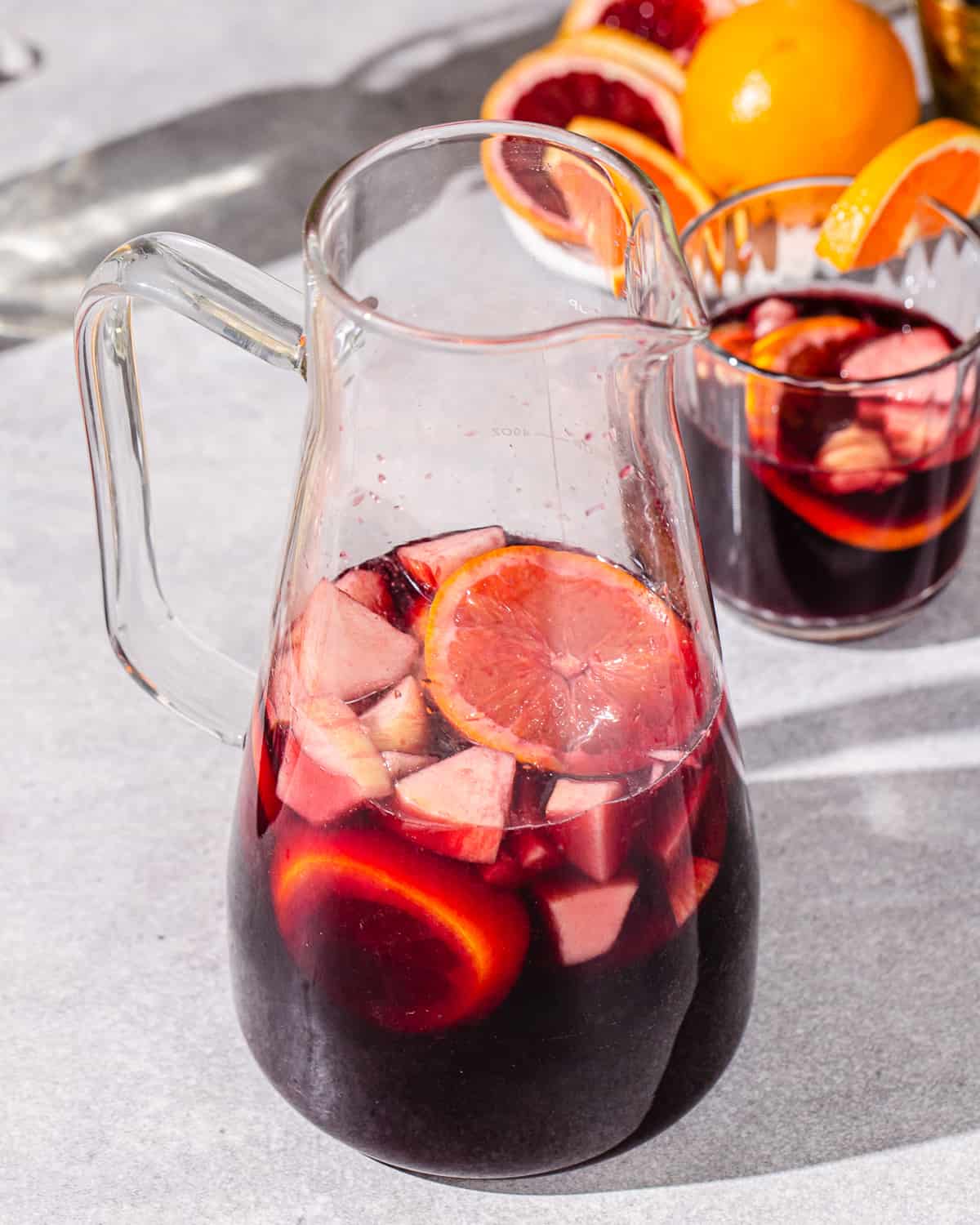 Glass pitcher full of non-alcoholic sangria on a gray countertop. A smaller glass of the sangria is on the right of the pitcher. The drink has cut up apples and orange slices floating in it.