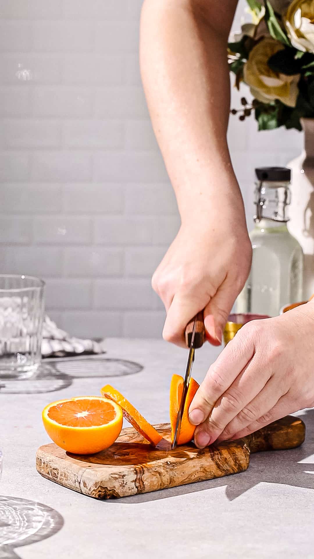 Hands cutting an orange into slices using a knife and cutting board.
