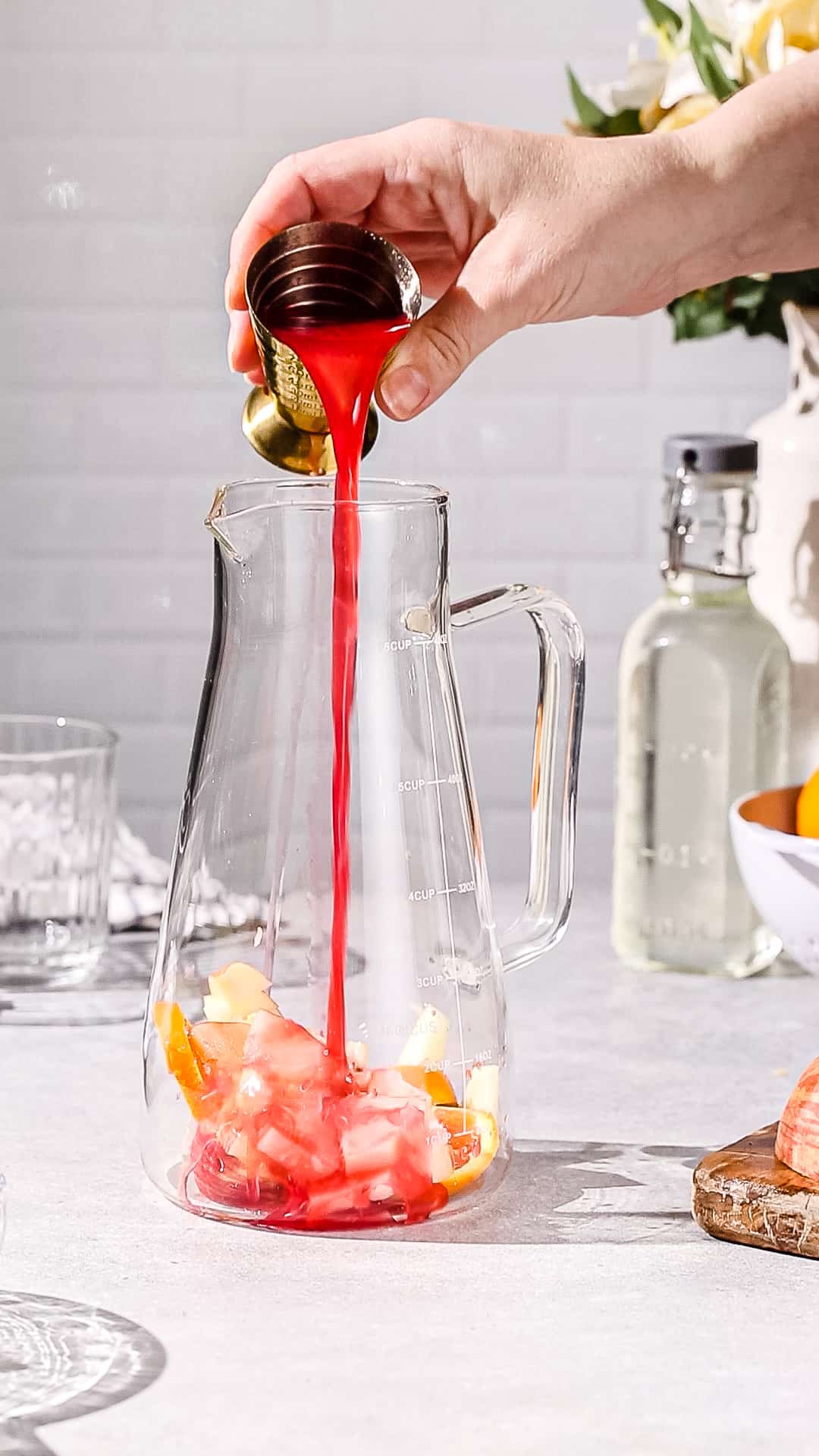 Hand using a large jigger to add a red colored juice to a glass pitcher that has cut up fruit at the bottom.