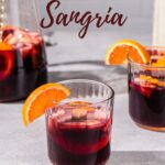 Semi-overhead view of a glass of non-alcoholic sangria. Apples and oranges are floating in the dark red drink and there is an orange slice on the rim of the glass. Another glass is off to the right and a pitcher of the sangria is to the left in the background. Text overlay at the top reads “Non-Alcoholic Sangria”.