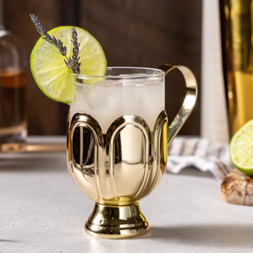 Side view of a Lavender Mule vodka cocktail on a gray countertop. The drink is in a gold and glass mug with a lime slice and dried lavender sprigs as garnish. In the background are a bottle of lavender syrup, some bar tools, and a cut lime on a wooden cutting board.