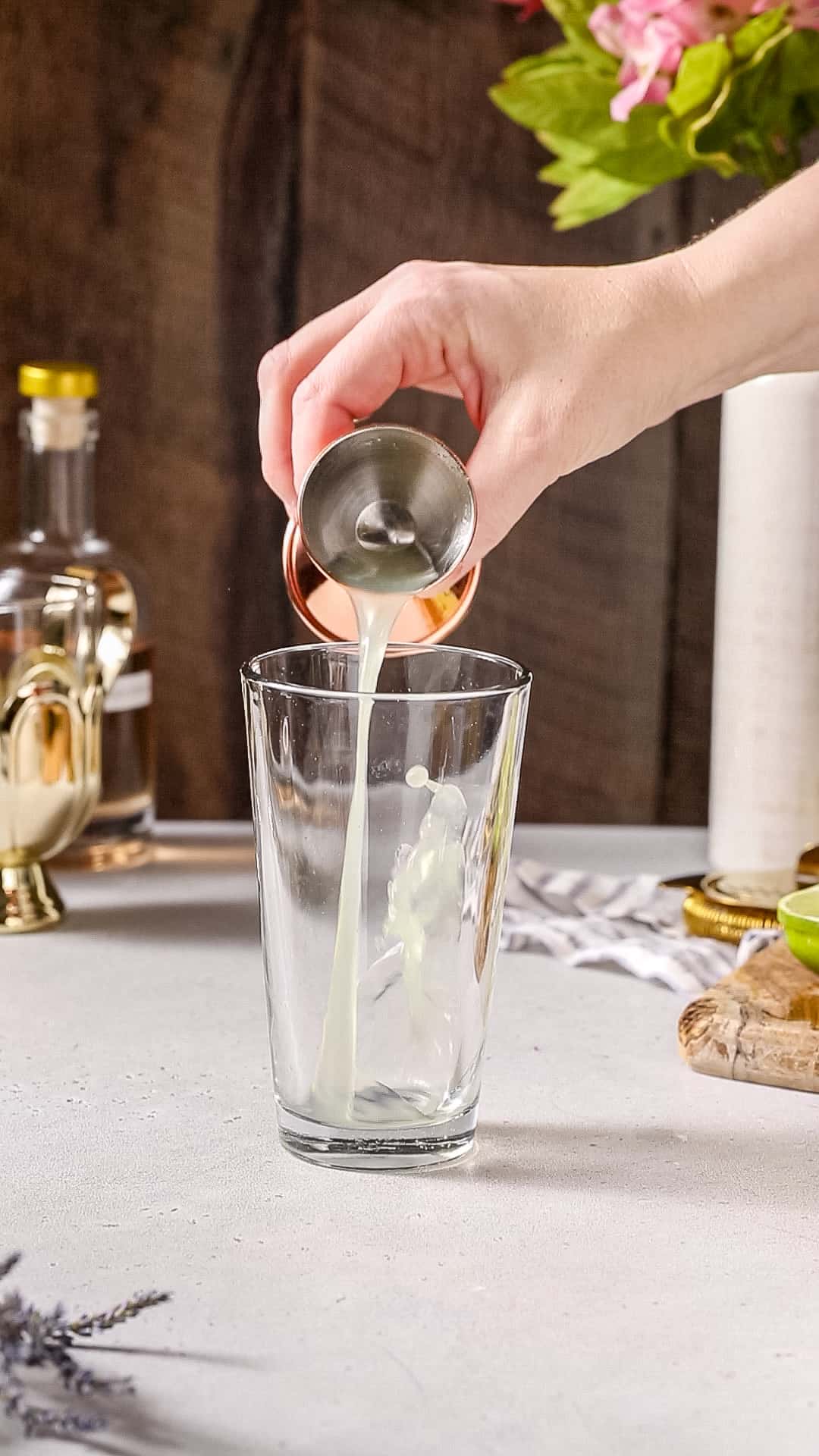Hand pouring lime juice from a jigger into a cocktail shaker.